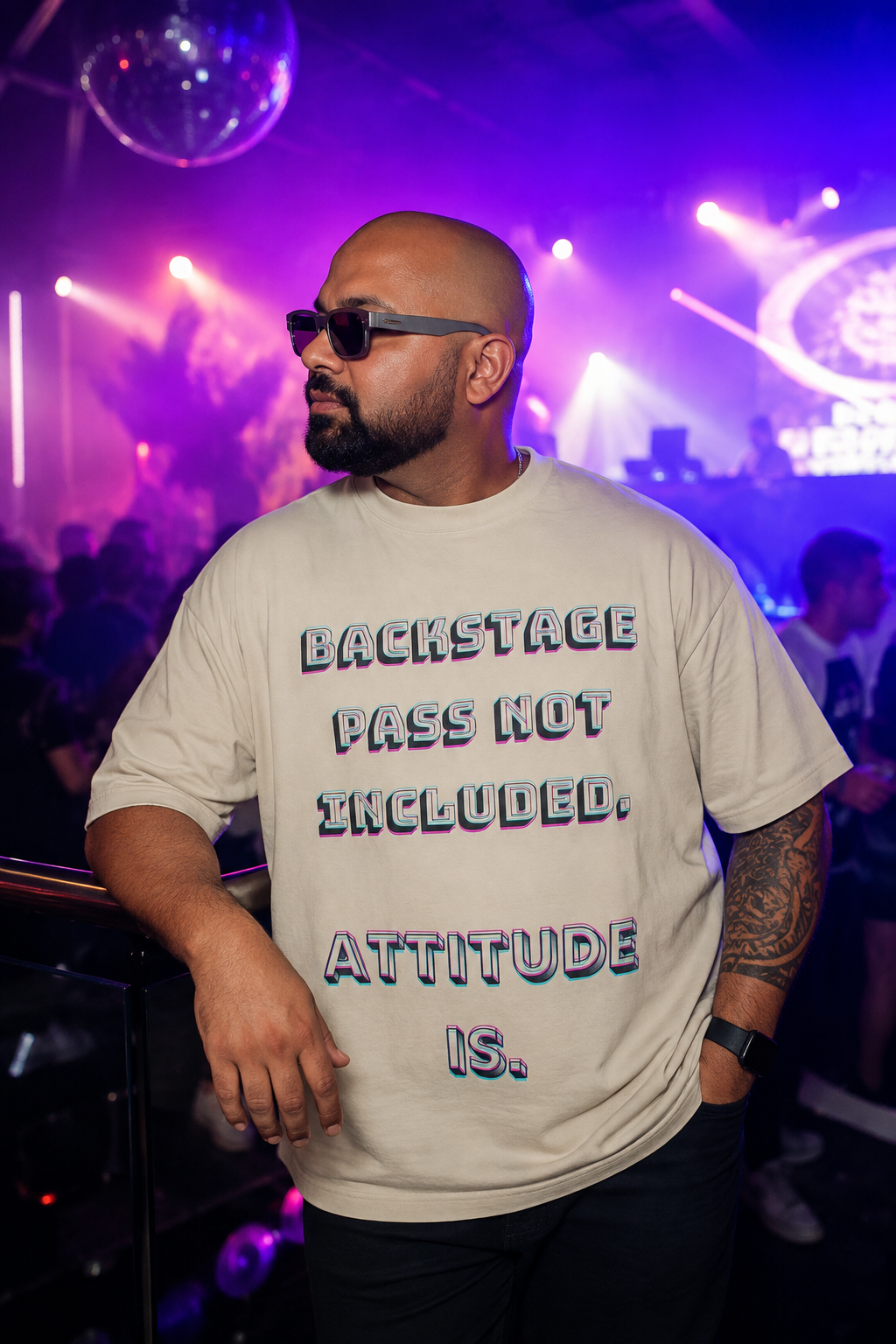 A man wearing a beige graphic t-shirt with the text 'BACKSTAGE PASS NOT INCLUDED. ATTITUDE IS.