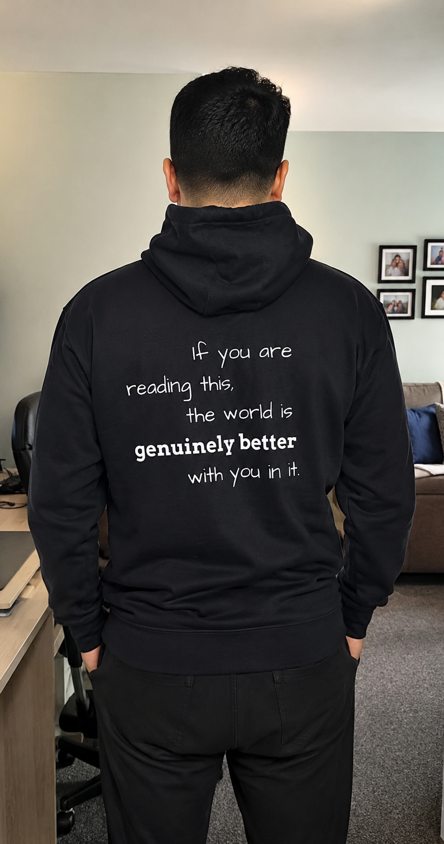 A man viewed from behind wearing a black hoodie with white printed text on the back that reads, 'If you are reading this, the world is genuinely better with you in it.' The photo is taken in an indoor room with a desk, chair, and couch visible in the background.