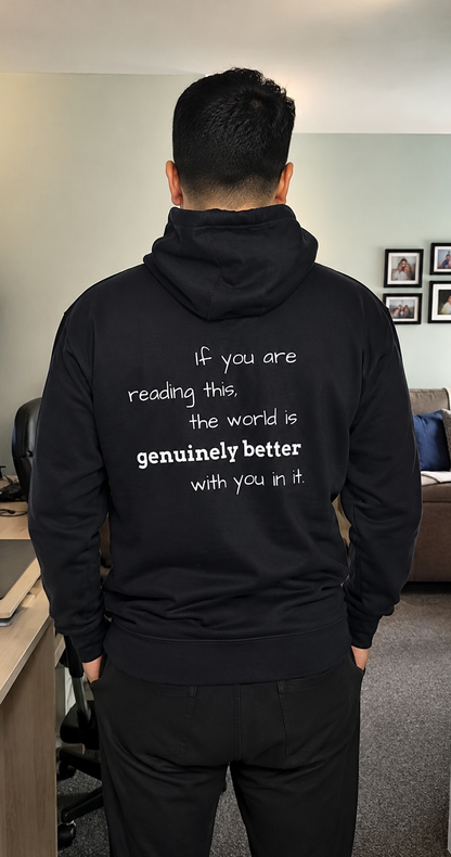 A man viewed from behind wearing a black hoodie with white printed text on the back that reads, 'If you are reading this, the world is genuinely better with you in it.' The photo is taken in an indoor room with a desk, chair, and couch visible in the background.